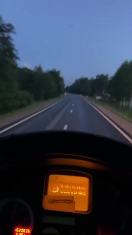 The video shows a first-person perspective from inside a vehicle, likely a truck, as it drives down a two-lane road during dusk. The dashboard is visible, with a bright orange digital display showing "Brake system truck warning." The road is wet, reflecting the dim light. Trees line both sides of the road, and there are occasional orange road markers. The sky is a deep blue, indicating the transition from day to night. The vehicle appears to be moving at a moderate speed.