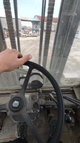 The view is from inside the cab of a forklift, looking out through the dusty windshield. A person's hand rests on the black steering wheel, and their legs and shoes are visible in the lower part of the frame. Through the windshield, stacks of wooden pallets can be seen in a yard or industrial area. In the background, there are buildings with corrugated metal roofs and a few people moving around. A rearview mirror shows the top of the forklift operator's head and shoulders. The cab interior looks aged and dusty.