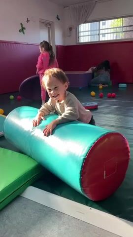 This video captures a lively scene in what appears to be an indoor children's soft play area or sensory room, likely located in Kiryat Bialik, Israel. The primary subject is a young child, a toddler with light brown hair, wearing a light-colored long-sleeved shirt and dark pants. The child is positioned on a large, teal-colored cylindrical soft play toy with red ends, actively pushing it forward or attempting to climb over it. The child looks towards the camera with a slight smile and engaged expression.

In the background, two other children are visible. One child in a pink outfit is seen moving across the room, while another child with dark hair is sitting on the floor further back, near a purple soft mat, seemingly interacting with small, colorful balls. The room features padded red walls, providing a safe environment, and a dark green mat covering the floor. Various other soft play items, including green rectangular mats and an assortment of red, yellow, and blue balls, are scattered throughout the space.

Bright natural light streams in from a large window on the right side of the frame, which has grates and light-colored curtains, indicating it is daytime. Butterfly decals are visible on the white wall in the upper left corner, adding a decorative touch. The overall atmosphere is bright, playful, and energetic. There is no discernible text visible within the frame.