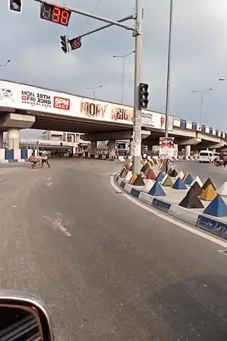 The video captures a street scene in a city, possibly in Nigeria, during daylight hours. The camera appears to be mounted inside a vehicle, likely a car. 

A major feature of the scene is an overpass, with large advertisements displayed on it. One advertisement prominently features the words "GLORY REIGNS" and another displays "Share a Coke." Traffic lights are visible, indicating a countdown with digital numbers.

Pedestrians and motorcycle riders are seen traversing the street. Some pedestrians are pushing carts. A red advertising pillar for "Kilimanjaro" is visible near a building with a large advertisement for "NOTE 50Pro," showcasing a mobile phone.

Traffic markers, in the shape of pyramids with alternating blue, white, and terracotta colors, are positioned along the road edge. A blue car is partially visible in the foreground. The sky is overcast, suggesting a cloudy day.