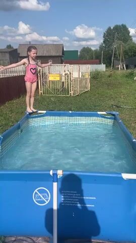 This video captures a young girl in a pink swimsuit jumping into a blue, above-ground swimming pool. The setting appears to be a backyard in a rural or semi-rural area, possibly in Nizhnii Bugrysh, Russia, given the context. Wooden houses and a fence are visible in the background, along with green grass and trees under a bright, sunny sky with some clouds, suggesting it is daytime and the weather is pleasant.

The girl, with her arms outstretched, leaps from the edge of the pool. She makes a splash as she enters the water, then resurfaces and settles into the pool. She appears to be enjoying herself, looking towards the camera with a smile.

The pool itself is rectangular with a blue liner and white frame. There is visible text on the side of the pool, which includes warnings in multiple languages: "NO SLOPING GROUND," "NE PAS FAIRE DE TERRAIN EN PENTE," "NO SOBRE LA PISCINA EN ZONA CON DESNIVEL," "KEIN NEIGENDER BODEN," "PAS VAN AFVLAKKENDE ONDERGROND," and "NIET OPZETTEN OP AFVLAKKENDE ONDERGROND." The text warns against setting up the pool on sloping ground. Another sign is visible in the background on a yellow gate, with Cyrillic text that reads "ОТВЕТСТВЕННОЕ ЛИЦО" which translates to "Responsible Person."