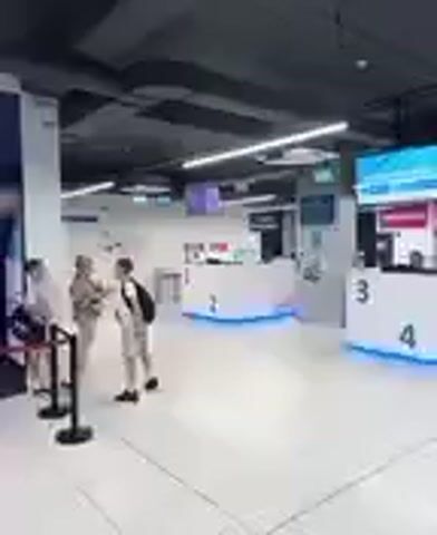 The video shows the interior of a modern, brightly lit building, likely a service center or a counter area in a public facility. The scene features several white, curved service counters, some numbered (2, 3, 4). The counters have blue LED lighting accents, and some have computer monitors and screens on them. Behind the counters, there are employees attending to customers or preparing for service.

In the foreground, three individuals are visible. Two are standing near a red stanchion and rope barrier, appearing to be conversing or waiting. One is wearing a light-colored outfit, and the other has on a backpack and light-colored pants. A third person, facing away from the camera, is also near the counters. The overall atmosphere seems calm and organized, suggesting a typical operational day.

The ceiling is dark with exposed beams and lighting fixtures, contributing to the contemporary design. Digital screens are mounted on the ceiling and walls, displaying information or advertisements, though the text is mostly illegible due to the video's resolution. The floor is a light-colored tile, reflecting the bright lights.

While no specific activity is clearly defined, the presence of service counters and people suggests transactional or informational exchanges are taking place or about to take place. The location context of Reda, Poland, indicates this is likely a public service point within that region. No specific time of day or weather can be deduced from the interior setting.