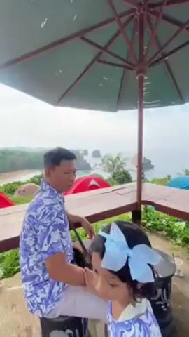 The video shows a man sitting at a wooden table under a large, open, green umbrella. He is wearing a blue and white floral shirt and white pants, and has a bag slung across his shoulder. A young child with a large blue bow in her hair is also visible in the foreground. The table overlooks a landscape of green foliage, tents (red and blue), a sandy beach, and the ocean with rock formations in the distance. The sky is overcast. The camera pans slightly to show more of the tents and ocean view.