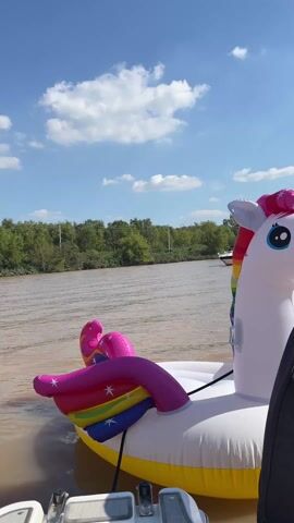 The video shows an inflatable unicorn floating on a river. The river is a muddy brown color and is surrounded by green trees. The sky is blue with scattered white clouds. The unicorn is white with a rainbow mane and tail and a yellow horn. It is attached to a boat, which is slightly visible in the lower-left corner of the frame. The camera angle is from behind a person, who is partially visible on the right side of the frame, looking towards the unicorn and the river. Another boat is seen in the background.