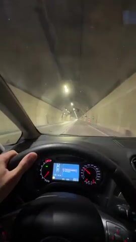 The video provides a first-person perspective from inside a car driving through a well-lit tunnel. A person's right hand is visible gripping the steering wheel. The car's dashboard is prominent in the foreground, featuring a speedometer and tachometer with red needles and backlighting. A central digital display on the dashboard shows "20°C" and "13:23", along with navigation information in Cyrillic script, indicating a remaining distance of "201.0 km" and an estimated travel time of "2:23".

The tunnel features arched concrete walls and ceiling, illuminated by numerous overhead fluorescent lights. Several large ventilation fans are visible overhead. Green lights are seen intermittently along the tunnel walls, possibly indicating emergency exits or safety features. Road markings and orange traffic cones are visible on the right side of the road. Other vehicles can be seen in the distance. An audible voice, likely from a GPS system, speaks in a Slavic language, asking "Do you want without unnecessary chatter?"

The setting is a road tunnel in Đurmani, Montenegro.