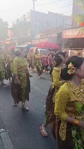 The video shows a group of young women in traditional Indonesian attire walking down a street. They are wearing brightly colored costumes, including green blouses adorned with gold embroidery and patterned skirts. Their hair is styled up, and some have flowers in their hair. They appear to be part of a procession or parade. The street is lined with shops and stalls, and there are other people in the background watching the procession. The lighting suggests it is daytime, possibly late afternoon, with a hazy or slightly overcast sky. The women are walking in formation, and some are looking towards the camera. The overall atmosphere seems festive and celebratory.