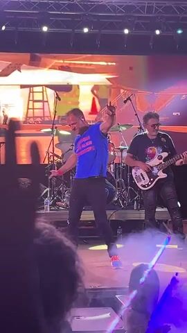 A male performer in a blue t-shirt and black pants is dancing on a stage in front of a large screen. The screen displays an image of a private jet parked on an tarmac, with a person in a large bear costume standing near it. Another performer, wearing a black shirt and jeans, is playing a bass guitar to the right of the main performer. Behind them, a drummer is visible behind a drum set. Smoke or fog is present on the stage, and stage lights are visible in the foreground. A crowd of people is in the foreground, out of focus.