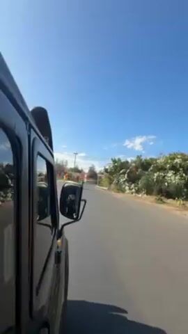 The video shows a view from inside a dark-colored vehicle, possibly a jeep, as it drives on a paved road. The camera is positioned to capture the side of the vehicle, including the side mirror and snorkel. The sky is bright blue with a few scattered clouds. The road is lined with vegetation, including bushes with white flowers. Other vehicles, including a scooter and a white car, can be seen moving on the road ahead. The overall environment suggests a sunny day in a semi-urban or rural setting.