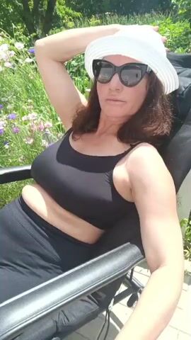 In this video, a woman is relaxing in an outdoor chair on a sunny day. She is wearing a white sun hat, black sunglasses, a black tank top, and black shorts. Her arm is resting on her head as she looks towards the camera with a slight smile. The background consists of lush green foliage and some wildflowers, suggesting a garden or park setting. The lighting indicates it is daytime, and the bright sunshine implies warm weather. The woman appears calm and content. There is no visible text in the video.