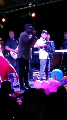 The video captures a live performance on a stage at night. A male singer, wearing glasses and a light-colored shirt, stands center stage, holding a microphone and gesturing to the crowd. To his left is a young woman in a red gown and a crown, presumably the guest of honor. Behind them, a band accompanies the singer, featuring a guitar player, an accordion player, and a keyboardist. The stage is decorated with colorful balloons. The scene appears to be a celebration, possibly a quinceañera, with an audience visible in the foreground. The lighting is a mix of spotlights and ambient stage lights.