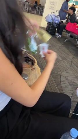 The video shows a young woman with long dark hair, wearing a white tank top and black pants. She is sitting in an airport waiting area, holding a bag of green peppers and a few packets of condiments. Her left arm is visible, and she has a rose tattoo on her upper arm. In the background, there are rows of seats, luggage, and other passengers. The woman is looking towards the camera and smiling. The scene appears to be taken during the daytime.