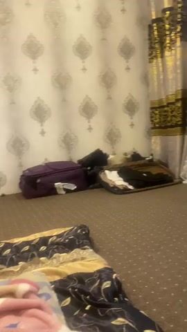 This media file depicts an indoor scene, likely a bedroom, with a focus on luggage and a bed. A purple suitcase is prominently displayed on the carpeted floor, next to a pile of clothing in a basket. The wallpaper features a repeating ornate pattern in silver and white. In the foreground, a neatly made bed is covered with a colorful blanket. To the left, a desk with a laptop and a bottle of water is visible. The lighting suggests it might be daytime, though the exact time is difficult to determine. There are no people visible in the frame. The overall impression is one of preparation for or aftermath of travel, with a sense of domesticity. There are no discernible location cues specific to Atkarsk, Russia.