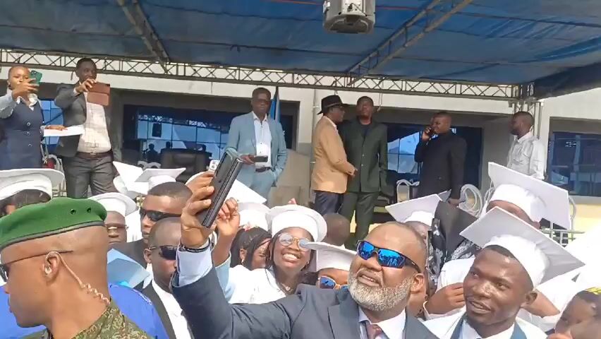 A crowd of people are visible in an outdoor setting. Most of them appear to be young graduates wearing white gowns and mortarboard hats. A middle-aged man in a navy blue suit and sunglasses is holding up his smartphone, seemingly taking a selfie with the group. The man has a grey beard and a patterned red tie. Several people are smiling and looking towards the camera. Some individuals in the background are wearing suits and other formal attire. There is a building visible in the background as well. A man in military uniform is partially visible on the left side of the frame.