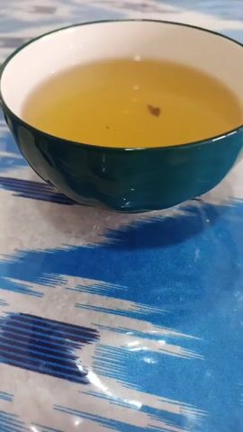 A low-angle, close-up video shows a bowl of liquid, likely tea, and a decorative glass jar containing tea or herbs on a table with a blue and white patterned tablecloth. The bowl is dark green on the outside and white on the inside, filled with a light yellow liquid that has a few small particles in it. The jar, which has a decorative gold top, sits further back on the table. A washing machine is partially visible in the blurry background.