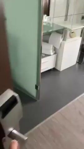 The video shows a modern, white hotel room. As the camera pans, we see a bed with white linens and a yellow throw, a desk with a white chair, a glass sink with towels and toiletries neatly arranged underneath. The room is bright and appears clean.