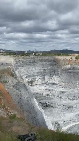 The video shows a large open-pit mine under a cloudy sky. The mine has multiple stepped levels with roads winding down to the bottom. Several yellow construction vehicles are visible within the mine. A few buildings and hills are visible in the distance on the horizon. In the foreground, some tires and green vegetation are visible. The camera slowly zooms out, providing a wider view of the mine.