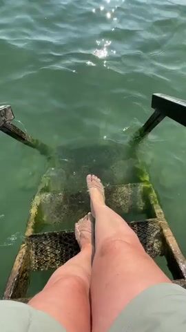 A first-person perspective video shows a person stepping down a submerged set of wooden stairs into clear, green water. Sunlight glints off the water's surface, creating a shimmering effect. The person's legs and feet are visible, with the water reaching just above their ankles. The stairs appear weathered and are covered in some algae. Two dark, wooden handrails extend from the top of the stairs into the water on either side. The overall impression is one of a serene, outdoor aquatic environment.