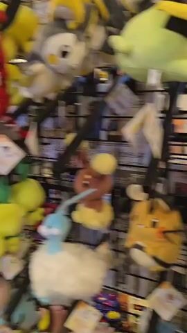 The video pans across a store filled with video game merchandise. Various plush toys, including characters from Super Mario and Pokemon, are displayed on shelves and racks. Several arcade cabinets are also visible, featuring games such as Star Wars: Return of the Jedi and an unidentifiable racing game. A glass display case showcases more merchandise, and large plush figures of characters like Jabba the Hutt and Pikachu are mounted on the wall above the arcade machines. The store appears well-lit and organized, with a carpeted floor.