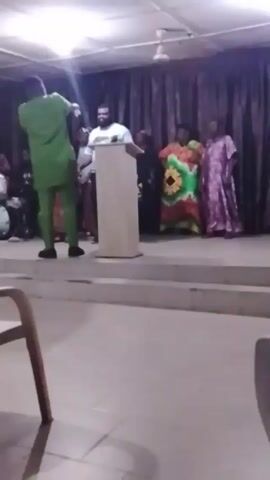 A man in a green suit stands in front of a white podium on a stage, speaking to an audience. Another man in a white t-shirt stands behind the podium. Two women in colorful dresses stand on the right side of the stage. There are drums visible in the background to the left. The room is lit with artificial light and the stage is covered in white linoleum. The camera is still, and a wooden chair is partially visible in the foreground.