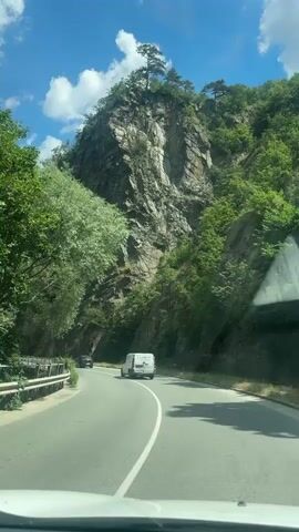 The video shows a white van driving on a winding road through a mountainous, forested area. The road is paved and has white lane markings. The mountains are covered in green trees, and there are rocky cliffs on the side of the road. The sky is blue with some white clouds. There is another car visible in the distance on the road. The video is shot from inside a vehicle, likely a car, looking forward through the windshield.