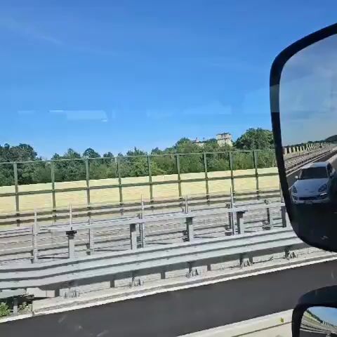 The video depicts a view from a moving vehicle, likely a train, traveling along a railway track. The scene is dominated by a beige and green noise barrier, beyond which is a lush, green hillside. On the hill, a building with a tower-like structure is visible amidst the trees. The sky is a clear, bright blue, suggesting a sunny day. A utility pole can be seen briefly at one point.  The focus remains on the passing landscape, highlighting the building on the hill.