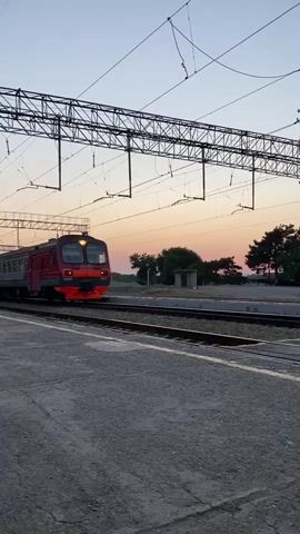 The video shows a train station at dusk. A red and gray passenger train is pulling away from the station. The train has multiple cars, and the windows are illuminated. The station platform is made of concrete, and there are train tracks visible. Overhead, there are electrical wires and support structures for the train's power system. In the background, there are trees and a few buildings. The sky is a pale orange and pink, indicating the sunset.