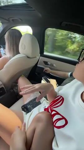 The video shows the interior of a car. Three young women are visible. The woman in the foreground is wearing a white top with red accents and white shorts with lace trim. She is sitting in the back seat, with her legs crossed and extended. A smartphone is visible on her lap, along with a white bag. Her right hand is raised, and she appears to be speaking or gesturing. Another woman is sitting next to her in the back seat, also with her legs extended. She is wearing a white top and is holding a phone. The third woman is in the driver's seat, facing forward and looking at her phone. The car's sunroof is open, and sunlight is streaming in. Trees and foliage are visible through the car windows, suggesting the car is in motion. The overall scene depicts a casual and lively atmosphere inside a moving vehicle.