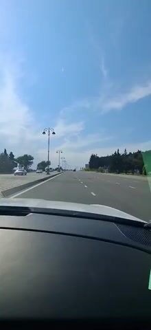 The video shows a view from the dashboard of a car driving down a multi-lane road on a clear, sunny day. The road is lined with trees and ornate lampposts. In the distance, more cars and trees are visible. The sky is a bright blue with some scattered white clouds. There are no people visible in the video, except those potentially inside the other cars. The overall mood is peaceful and relaxed, suggesting a pleasant drive in an urban or suburban area. The location is identified as Khatay Raion, Azerbaijan.