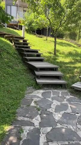 The video shows a wooden staircase and pathway leading up a grassy slope towards several modern buildings. The staircase is made of weathered gray wood, with each step supported by dark wooden beams. Some of the steps appear to have integrated LED lighting, which is faintly visible. The pathway in the foreground is paved with large, irregularly shaped gray flagstones. Lush green grass covers the hillside, and trees with full green foliage are present, casting shadows on the ground. In the background, there are contemporary structures with gray siding and large windows, suggesting a residential or resort setting. The overall scene depicts an outdoor environment with a focus on natural landscaping and modern architecture.