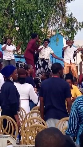 The video captures an outdoor performance with a group of people on a raised platform, likely a stage. A male performer in a maroon suit is center stage, singing into a microphone and gesturing with his hands. To his right, a female performer in a white top and patterned skirt is also singing and dancing. To the male performer's left, another male performer in a white shirt and patterned pants sings into a microphone. Behind them, other individuals are visible, including someone playing a saxophone and others singing. In the foreground, an audience is gathered, seated on chairs and standing. The environment appears to be a public gathering with trees and tents in the background. The atmosphere seems lively and engaging, suggesting a musical event or celebration.