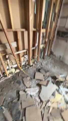 The video begins with a shaky, out-of-focus shot of a room under construction. Studs are visible, along with exposed electrical wiring and plumbing. Debris covers the floor. The camera then pans to a person holding a camera, and then to a man climbing a ladder in a narrow hallway. The hallway walls are white, and the ceiling is exposed with ductwork. The video returns to the construction site, showing more of the unfinished room, including an open doorway and more exposed studs. The scene appears to be a residential or commercial space undergoing renovation.