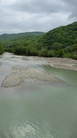 The video shows a wide, slow-moving river with grey water, multiple gravel bars, and green forested hills in the background under an overcast sky. The camera seems to be positioned on a bridge overlooking the river. There are no people or animals visible in the video.