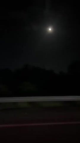 The video shows a car driving on a road at night. A bright moon is visible in the sky. Trees line the side of the road, silhouetted against the night sky. The guardrail along the road is white. Another vehicle passes by.