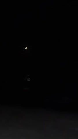 The video shows a dark, grainy scene of a crescent moon hanging in the night sky above a body of water. The moon is a pale yellow sliver, and its reflection shimmers on the dark surface of the water. The lower part of the frame shows a dark, textured surface, possibly sand or a beach, with faint, almost indiscernible lighter patches. The overall impression is one of a very dark, calm night, likely by the sea, with minimal light from the moon.