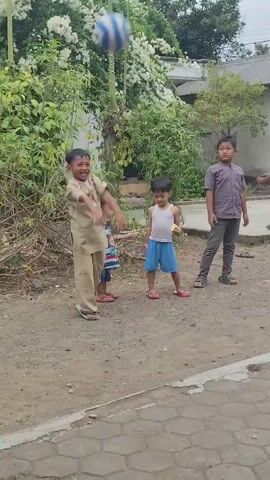 A group of children are playing with a ball outdoors. The scene appears to be in a rural or suburban area, with a dirt ground and some buildings in the background. The children are of various ages, and they are engaged in playing with a striped ball. Some of the children are kicking the ball, while others are watching or waiting for their turn. The weather appears to be overcast.