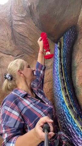 A blonde woman in a plaid shirt holds a red garland up to a colorful, scaly, serpent-like sculpture. She is holding a selfie stick in her right hand, and the garland in her left. The sculpture is against a textured brown rock wall. The woman smiles at the camera.