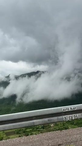 The video shows a view from a roadside, focusing on a metal guardrail with an advertisement sticker on it. Beyond the guardrail, there are green trees and foliage. In the background, there are mountains partially obscured by heavy, low-hanging clouds, creating a foggy atmosphere. The sky is overcast and gray. The camera seems to be moving forward, possibly from a vehicle.