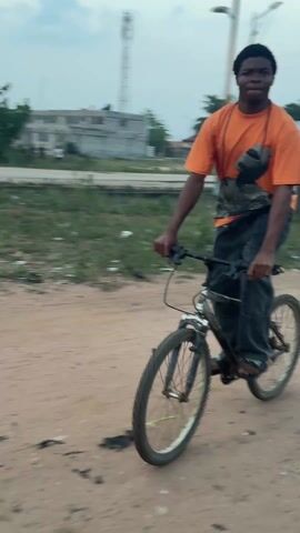 A young man rides a bicycle down a dirt road. He is wearing a bright orange t-shirt with a graphic print, dark trousers, and sandals. He has a crossbody bag slung over his shoulder. The bicycle is old and appears to be slightly rusty. The road is unpaved and sandy, with patches of grass and debris scattered along the sides. In the background, there are trees, buildings, and a tall tower structure. The sky is overcast, suggesting it is early morning or late afternoon. The video follows the cyclist as he moves forward, with the camera occasionally blurring due to the movement.