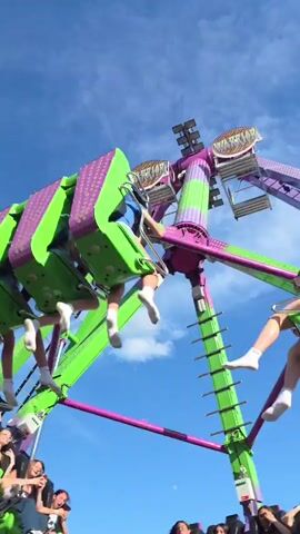 The video shows a large amusement park ride, possibly a pendulum ride or a similar thrill ride. The ride features several gondolas or cars, each holding multiple people, suspended from a central arm. The gondolas appear to be bright green with purple accents and have pink LED lights on the front. The ride is in motion, with the gondolas swinging and rotating against a clear blue sky with some scattered white clouds. People are visible in the gondolas, with their legs often hanging out or extended. Some people have their arms raised in the air. The ride is positioned in an outdoor setting, with the background showing a building with a light-colored facade and a fence. The overall impression is one of excitement and fun as people experience the thrill of the ride.