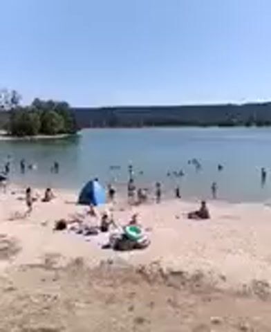 The video shows a sandy beach with a body of water. There are many people on the beach and in the water. Some people are sunbathing, others are swimming or playing in the water. There is a blue tent on the beach, as well as several beach towels and other items. The water is calm and the sky is clear. The overall scene is one of a busy summer day at the beach.