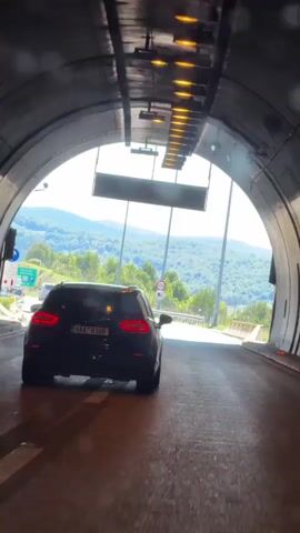 The video begins inside a dark tunnel, looking out towards a brightly lit exit. A dark colored SUV is driving out of the tunnel. The exit reveals a sunny day with a clear blue sky, rolling green hills, and trees. The road curves to the left, and there are streetlights and traffic signs visible. As the SUV continues to drive, the camera follows it out of the tunnel and onto the open road. The SUV passes a green sign with white text, and then a speed limit sign indicating 100. The video continues to show the SUV driving on the highway.