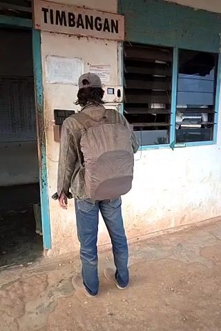 A person wearing a backpack and a baseball cap stands with their back to the camera, interacting with a machine mounted on a wall beneath a sign that reads "TIMBANGAN." The machine appears to be a ticket dispenser or a payment terminal. The person is wearing a grey jacket, blue jeans, and a backpack. To the right of the machine, a window with horizontal blinds is visible. A blue and white sign with an icon is affixed to the wall to the right of the window. Another person wearing a grey hooded sweatshirt, dark pants, and green boots walks into the frame from the right, approaches the first person, and appears to speak to them. The setting is outdoors, with a weathered building and a dirt ground. In the background, motorcycles, a truck, palm trees, and other buildings are visible.