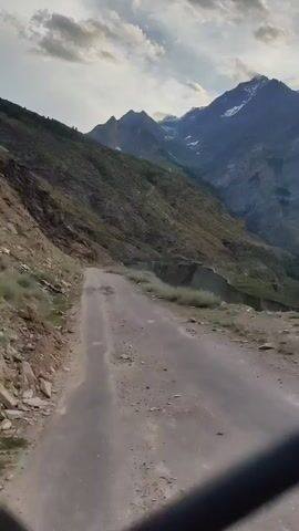 This is a first-person perspective video of a rough, unpaved road winding through a mountainous landscape. The road is dusty and appears to be in a state of disrepair with potholes and loose gravel. The surrounding terrain is steep and rocky, covered in sparse vegetation. In the distance, majestic, snow-capped mountains rise against a cloudy sky. The overall impression is one of a remote and rugged wilderness. The lighting suggests it is daytime, likely late afternoon or early evening, given the muted colors and cloudy conditions. There is no visible text in the video. The scene evokes a sense of adventure and the challenges of travel in a remote, high-altitude region.