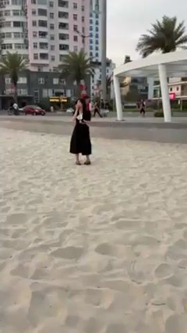 The video shows a young woman and a young man standing on a sandy beach. The woman is wearing a black dress, and the man is wearing a black shirt and black pants with a white stripe down the side. The woman raises her hand to her face and appears to yawn or cough. The man is looking at his phone. In the background, there is a busy street with cars, motorcycles, and people walking and biking. There is a building with a "COFFEE TIME" sign on it and a large white structure with pillars. Palm trees are visible throughout the scene, and the sky is overcast.