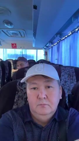 The video shows the interior of a bus with rows of seats. The camera is positioned in the front of the bus, facing backwards. A man wearing a white baseball cap and a dark patterned shirt is in the foreground, looking directly at the camera. Behind him, two other men are visible in the seats. The bus has blue curtains covering the windows. The lighting is dim and blue.