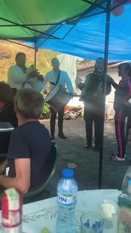 Four musicians are performing under a blue and green canopy for a small group of seated people. A man on the left is playing bagpipes, another in the center is playing a drum, and one on the right is playing an accordion and singing into a microphone. All three musicians are wearing white shirts and dark pants. A woman in a dark track suit with pink stripes stands beside the musician playing the accordion. Several people sit at a table in the foreground, watching the performance. On the table is a bottle of water, a glass, and a red beverage carton. The environment appears to be an outdoor gathering or small party, possibly in a backyard or patio.