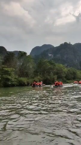 The video shows a group of people wearing orange life vests floating down a river on rafts. There are several rafts in a line. The people on the rafts appear to be paddling. The river is surrounded by green trees and forested hills, with a cloudy, overcast sky overhead.