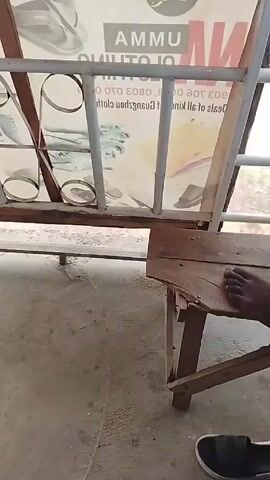 The camera shows a close-up shot of a cement floor, a wooden chair, and a portion of a sign. The sign appears to have an advertisement for clothing and some blurry images. The chair is simple and looks worn. A dark-skinned person's foot is visible on the chair. A sneaker lies on the cement floor. The scene seems to be outside or in a semi-outdoor space.