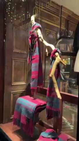 The video shows a store window display with scarves, a hat, and decorations. The display includes a wooden branch with scarves draped on it, a stack of scarves on a decorative base, and a hat resting at the bottom. The scarves are a blend of red, gray, and green colors. The backdrop of the display is designed to resemble wood paneling and ornate details. To the right of the scarves, other clothing items are visible on mannequins, along with some strings of lights. A quote is written in gold letters on the glass, but it is not fully visible.