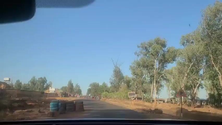 A car is driving down a road in a rural area. The road is paved and has dirt shoulders on either side. There are buildings and trees on both sides of the road. There are people walking on the side of the road. The sky is blue.